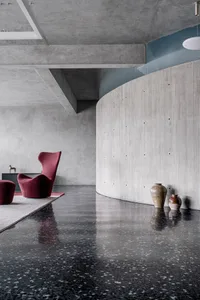 A minimalist living room with concrete walls, a red armchair, and terrazzo flooring.