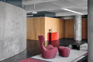 A modern lounge area featuring concrete walls and wood paneling, with a large burgundy chair and ottoman on a patterned rug.