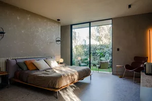 A minimalist bedroom with concrete walls, a wooden bed, and a view of a garden through large sliding glass doors.
