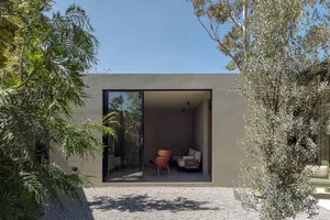 A modern residential exterior with concrete walls, large glass sliding doors, and a gravel courtyard surrounded by greenery.