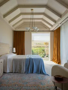 A minimalist bedroom featuring a bed with textured fabric bedding, a patterned rug, and large windows with yellow curtains, l
