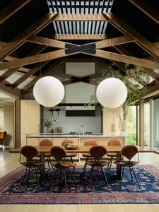 A modern dining room with a long wooden table surrounded by chairs, large pendant lights, and a blue rug with intricate patte
