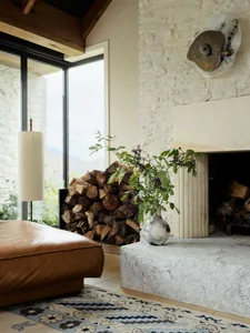 A stone fireplace in a living room with wood logs and a vase of greenery on the mantle.