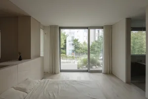 A minimalist bedroom featuring neutral colors, a large window with white curtains, and a view of trees and buildings outside.