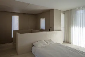 A sleek, contemporary bedroom with minimalist wooden paneling and a bed, featuring white bedding and sheer curtains.