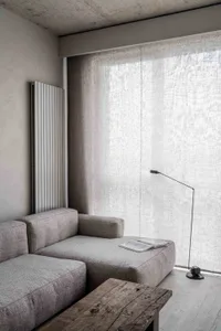 A minimalist living room with a light-colored fabric sofa, concrete radiator, cotton curtains, and a wooden coffee table.