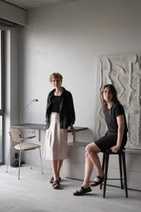 Two women standing and sitting in a minimalist room with white walls, wooden floor, and natural light.