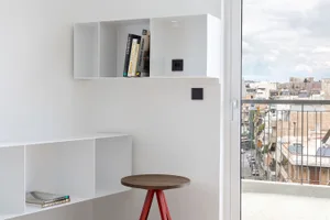 A minimalist office space with a white-painted wall, a wooden stool with a red metal frame, and a bookshelf with books