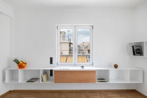 A minimalist residential interior featuring white walls, a large open window with a view of neighboring buildings, and wooden