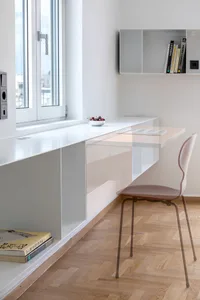 A minimalist workspace featuring a white desk with a single book and a bowl of fruits on top.