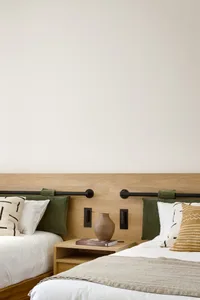 Minimalist bedroom features a bed with a wooden headboard, beige and white bedding, and a side table with a beige vase and bo