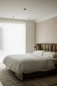 A modern bedroom with a brown leather bed, white blinds covering a window, and a white blanket on the bed.