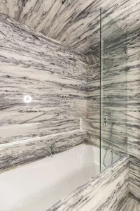 Modern bathroom with marble walls and floor, featuring a white bathtub and glass shower enclosure.