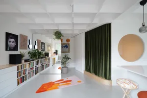 A minimalist living room with white walls, a white floor, and a mix of materials including green curtains, orange rug, and wo