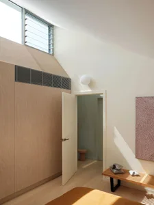 A modern bedroom with a tall, white wall featuring a large window and a wooden paneled wall with a door leading to another ro