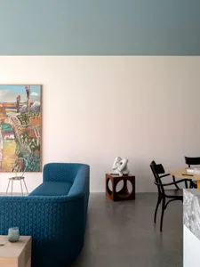 A modern living room featuring a blue couch, wooden stool, and abstract sculpture in front of a colorful painting.