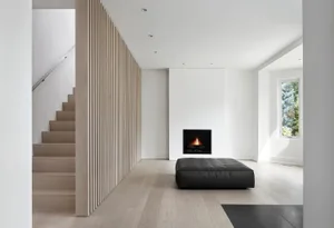 A minimalist living room featuring a light wood floor, a small black ottoman in front of a fireplace, and a white wall.