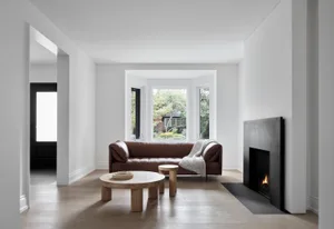 A minimalist living room featuring a brown leather sofa, wooden coffee tables, and a modern fireplace with a cozy blanket.