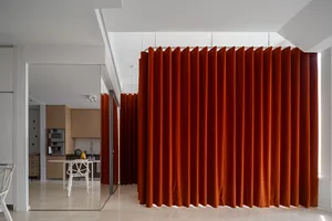 A modern apartment with bright orange velvet curtains in the foreground, white walls, and a kitchen in the background.