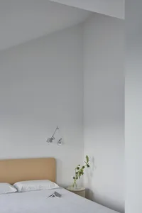 A modern minimalist bedroom featuring a bed with a fabric headboard and white bedding, placed in front of a white wall with a