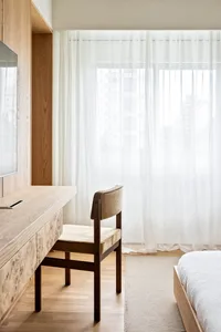 A modern minimalist bedroom with a wooden floor, a white bed with a light brown wooden frame, and a desk with a television.