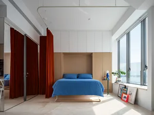 A modern minimalist bedroom with a blue bedspread, wooden headboard, and orange curtains. The room features a large window an