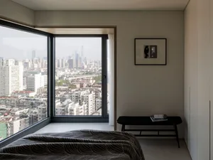 A minimalist bedroom with a bed, a black bench, and a cityscape view through the large window.