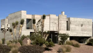 Modern, minimalist concrete house exterior with palm trees and native vegetation in the foreground.