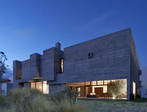 A modern house with a concrete exterior at dusk, featuring large windows and outdoor lighting.