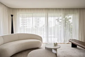 A modern living room with a white curved sofa, a marble coffee table, sheer curtains, and a large window.