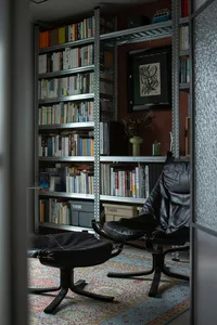 A modern library featuring metal shelves filled with books, a black leather chair and ottoman, and a patterned rug.