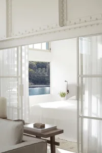 A minimalist bedroom featuring white textiles and a serene view of water through the window.