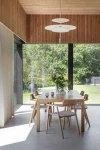 A modern dining room with wooden furniture and large windows overlooking greenery.