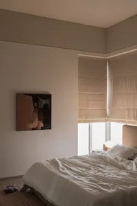 A minimalist bedroom featuring a bed with a cotton blanket, a painting on the wall, and light streaming through the window wi