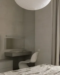 A minimalist bedroom with a bed, chair, desk, and mirror against a textured gray wall.