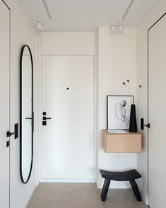 A modern, minimalist entrance hall featuring a white door with black handles, a floating wooden drawer with a black vase, and
