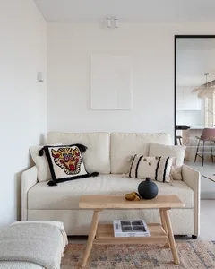 A minimalist living room featuring a neutral-toned sofa with decorative pillows, a wooden coffee table, and a rug, with a sim