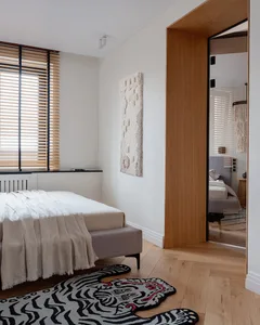 A minimalist bedroom featuring a bed with light wood frame and white bedding, surrounded by light wood floors and walls, and