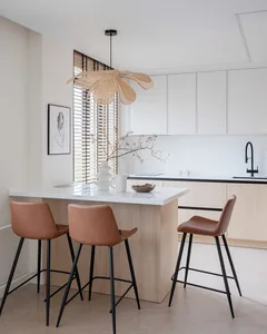 A modern minimalist kitchen with wooden finishes, a hanging woven ceiling lamp, and brown leather bar stools.