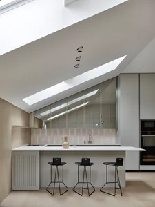 A modern residential kitchen with white and light-colored finishes, featuring skylights, a minimalist island, and black stool