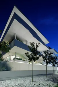 A modern white building with geometric architecture viewed at night with illuminated interiors and landscaping.