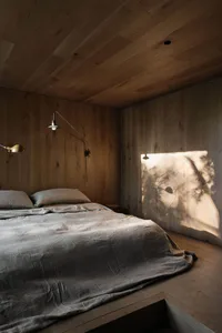 A minimalist bedroom with wooden walls and a bed with grey sheets, illuminated by natural light casting shadows on the wall.