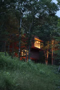 A modern wooden house nestled in a forested area with warm interior lights visible through large windows.
