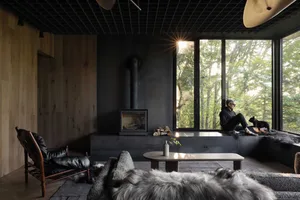 A modern minimalist living room featuring exposed concrete walls, large windows overlooking a forest, a fireplace, and a pers