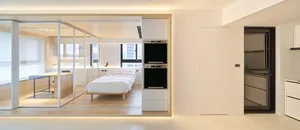 A modern, minimalist kitchen with white cabinets, a long wooden table, and a view of a bed and cityscape outside.