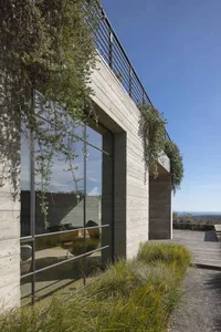A modern house with a concrete exterior and large windows, surrounded by greenery and a clear blue sky.