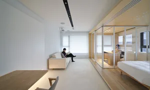 A modern living room with a white sofa and wooden flooring, separated from an office space with glass walls.