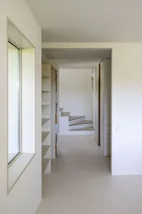A white concrete staircase leading from a minimalist hallway with a bookshelf and a large window.