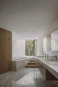 A minimalist bathroom with a marble floor, walls, and countertop, featuring a bathtub and a window with a view of trees.