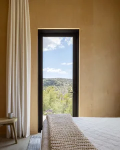 A minimalist bedroom featuring a bed with a brown wooden headboard, a small wooden stool with a mug, and a view of the outdoo